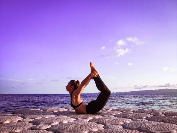 Avrupa görünümlü sportif bir kız deniz kenarındaki bir rıhtımda temiz havada yoga yapıyor.