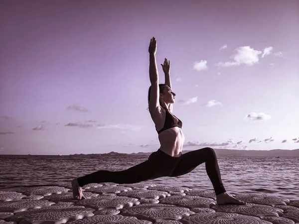 Avrupa görünümlü sportif bir kız deniz kenarındaki bir rıhtımda temiz havada yoga yapıyor.