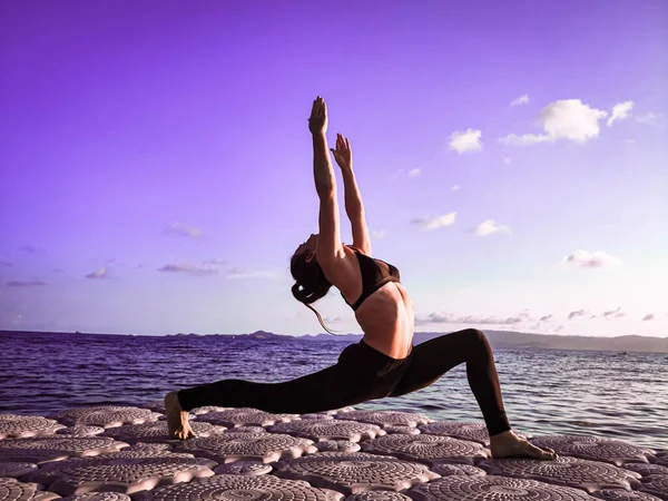 Avrupa görünümlü sportif bir kız deniz kenarındaki bir rıhtımda temiz havada yoga yapıyor.