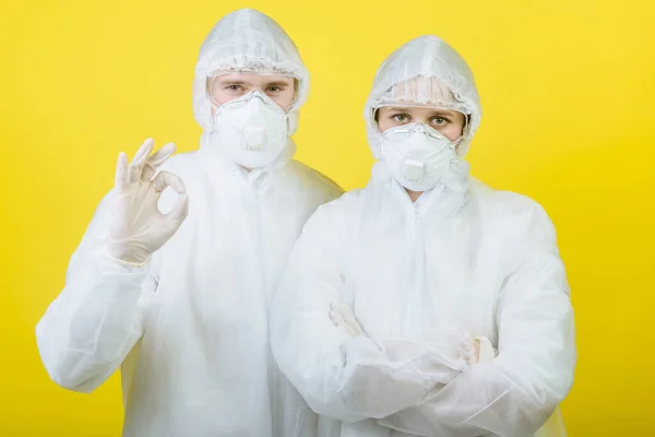 Two men in protective suit respirator masks isolated on a yellow background studio. The epidemic pandemic of the new rapidly spreading coronavirus 2019 ncov. COVID-19. Showing ok sign