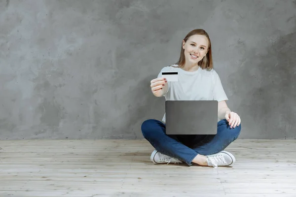 Happy woman sits on the floor on a gray background, shopping online with a credit card and computer. Online store