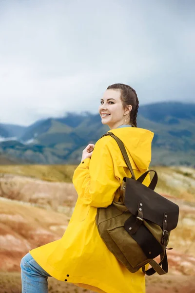happy woman traveler in a yellow cloak on a background of colored mountains. Travel and adventure concept