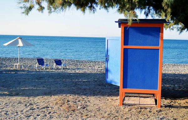 Açık kapısı, güneşliği ve mavi denizin yanındaki kumlu kumsalda iki boş plaj yatağı olan bir soyunma kabini, Yunanistan 'ın Girit adasındaki yaz tatillerinde güneşli bir günün daha sona erişini simgeliyor..