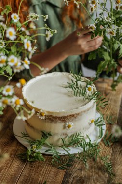 Tahta bir masanın üzerinde papatya çiçekleri ve yeşillik filizleriyle süslenmiş bir pasta var..