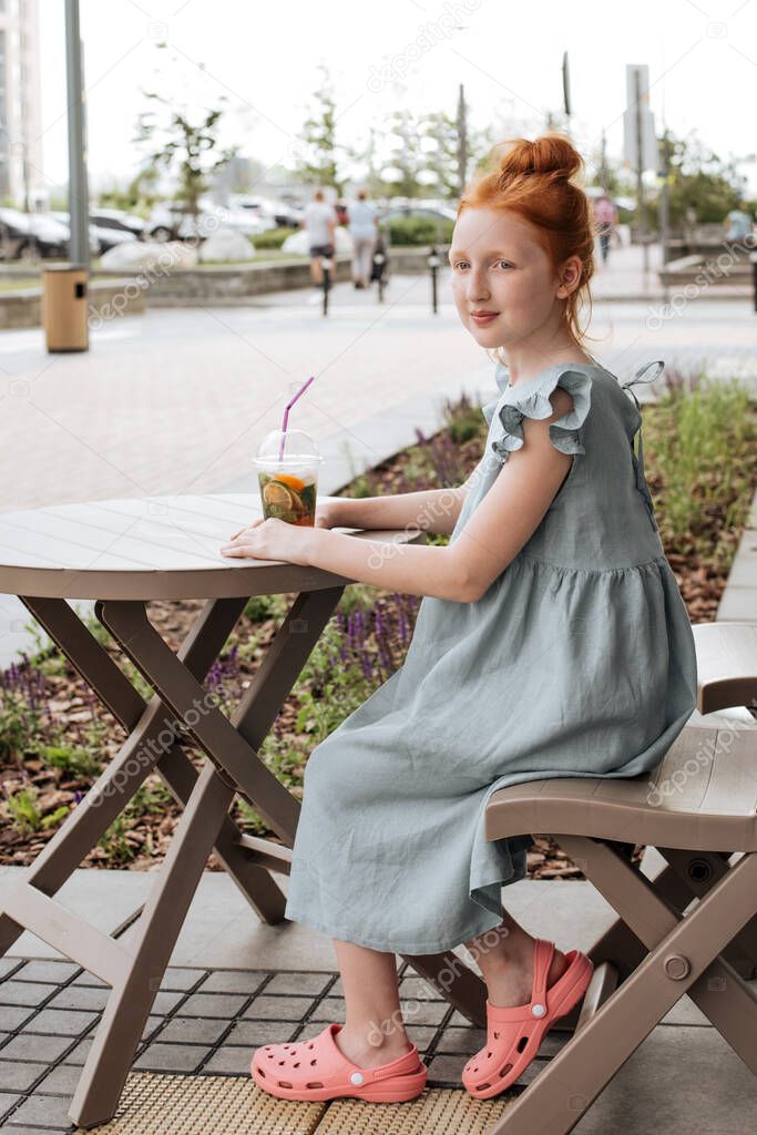 Red-haired girl sitting on a summer cafe with a glass of lemonade.