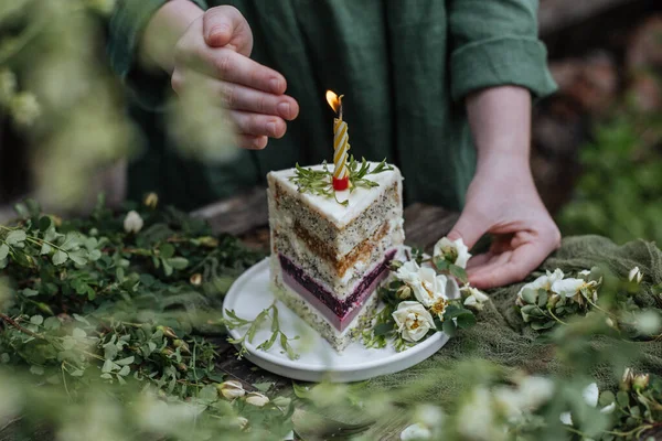 In the garden among the greenery on a wooden table a piece of birthday cake with a burning candle.