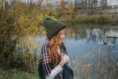 Örgü şapkalı kızıl kız ılık sonbahar gününde göl manzarasının tadını çıkarıyor..