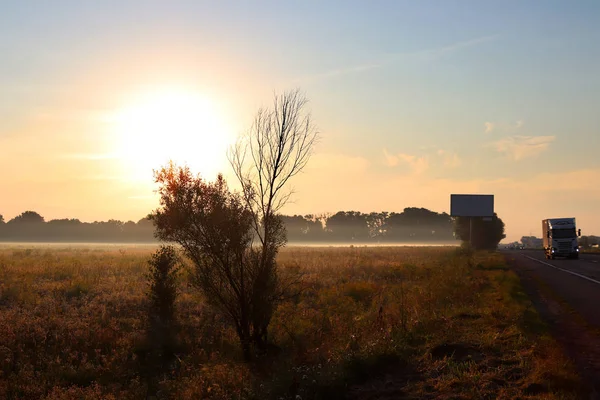 Dawn outside the city. Autumn begins. A huge sun rises over the yellowed grass. Morning fog. On the road there are cars and trucks