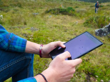 Adamın biri elinde tabletle çim alanda oturuyor.