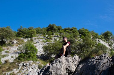 Genç yürüyüşçü kayada oturuyor, dinleniyor, dağlara bakıyor ve güzel manzaraya bakıyor.