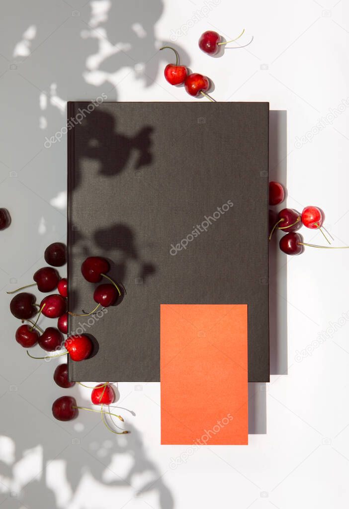 Orange business card and black folder with cherries under the shadows of the summer sun on a white background
