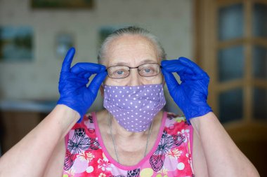 Koruyucu maskeli yaşlı, gri saçlı kadın lastik eldivenli gözlükleri ayarlıyor.
