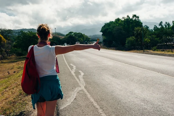 道路上の若いヒッピーの女の子ヒッチハイク。後景 — ストック写真