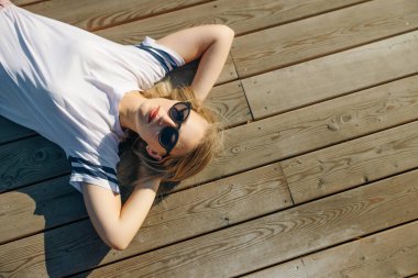 Hippi genç kadın ahşap zeminde yatıyor. Kot pantolon ve güneş gözlüklü beyaz bir tişört giy..