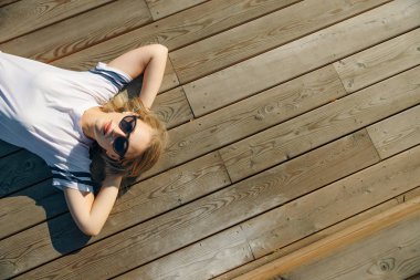 Hippi genç kadın ahşap zeminde yatıyor. Kot pantolon ve güneş gözlüklü beyaz bir tişört giy..