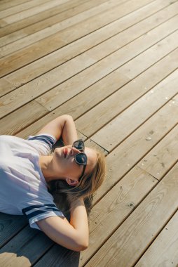Hippi genç kadın ahşap zeminde yatıyor. Kot pantolon ve güneş gözlüklü beyaz bir tişört giy..