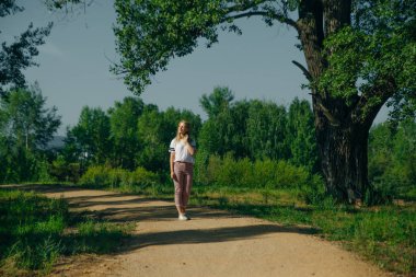 Hippi genç bayan yeşil bir parkta duruyor. Kot pantolon ve beyaz tişört giy.