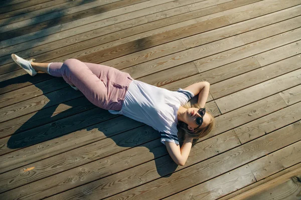 Hipsterska młoda kobieta leży na drewnianej podłodze. Noś dżinsy i biały T-shirt w okularach przeciwsłonecznych. — Zdjęcie stockowe