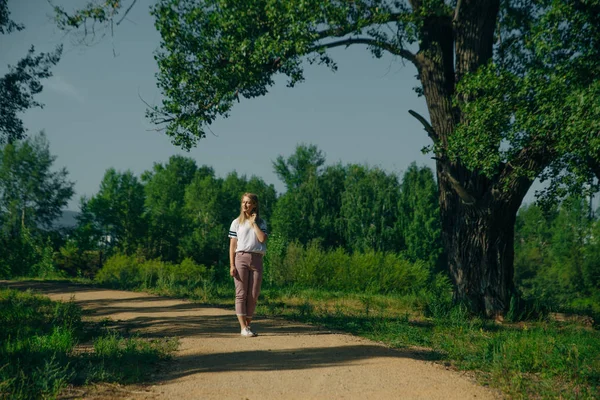 Hipster fiatal nő áll egy zöld parkban. Viselj farmert és fehér pólót. — Stock Fotó