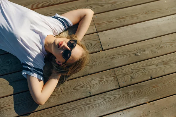 Хипстерская молодая женщина лежит на деревянном полу. Носите джинсы и белую футболку в солнечных очках . — стоковое фото