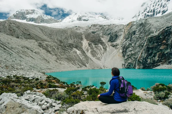 Laguna 69，秘鲁。 一个女孩背靠着蓝色的湖面坐着，穿着一件黄色的牛仔裤夹克 — 图库照片
