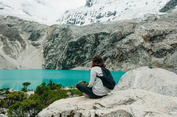 Laguna 69, Peru. Uma menina senta-se com as costas contra um lago azul em uma jaqueta amarela em jeans — Fotografia de Stock