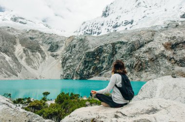 Laguna 69, Peru. Sırtı mavi bir göle yaslanmış, kot pantolonlu sarı ceketli bir kız oturuyor.