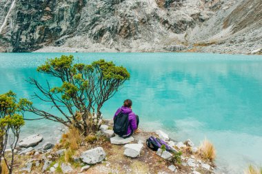 Laguna 69, Peru. Sırtı mavi bir göle yaslanmış, kot pantolonlu sarı ceketli bir kız oturuyor.