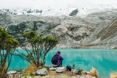 Laguna 69, Peru. Sırtı mavi bir göle yaslanmış, kot pantolonlu sarı ceketli bir kız oturuyor.