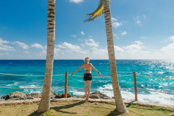 girl stands with her back against the blue ocean between palm trees. Cancun Mexico.
