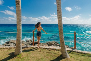 Kız sırtı mavi okyanusa dönük, palmiye ağaçları arasında duruyor. Cancun Meksika.