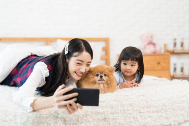 Hayvan sevgisi konsepti. Yaşlı kız kardeş ve kız kardeş kadın selfie Pomeranian Spitz köpekleri ve cep telefonu. Asyalı bir kız, genç bir kadın ve yavru bir köpek yatak odasında yatmayı dört gözle bekliyor..