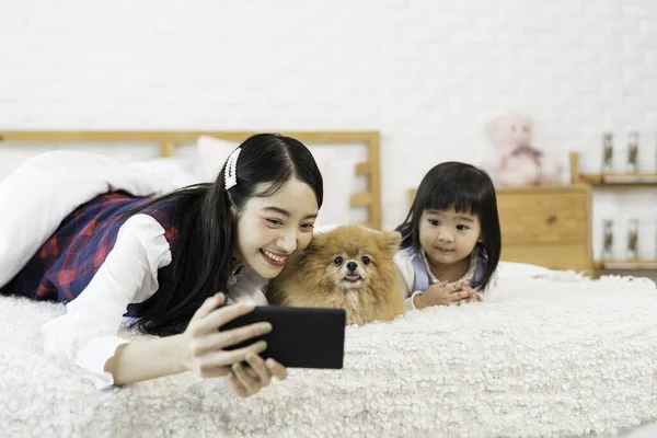 ペット愛好家の概念 姉と妹の女性が携帯電話でポメラニアン スピッツの犬を自撮り アジアの女の子と若いです女性と子犬楽しみますベッドルームでベッドの上に座って — ストック写真