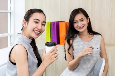 Kahve dükkanında oturan iki mutlu kadın cep telefonu ve tablet kullanıyor. Tablet kullanarak gülümseyen iki genç bayan. Sosyal ağı kullanarak tabletle çalışan bir kadın. Asyalılar. Alışveriş çantası..