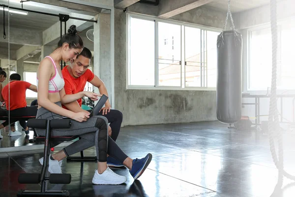 Female are discussing fitness with trainers. Young female talking during workout in gym. Woman coach explaining directives with laptop or computer.