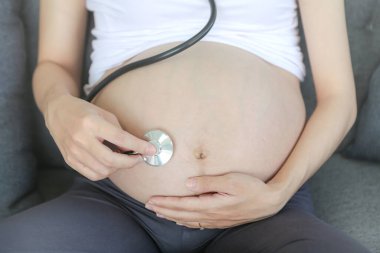 Anne steteskop kullanıyor doktor karnındaki bebeğin sesini dinliyor. Kanepede bebeğini dinlerken steteskop kullanan Asyalı hamile kadın..