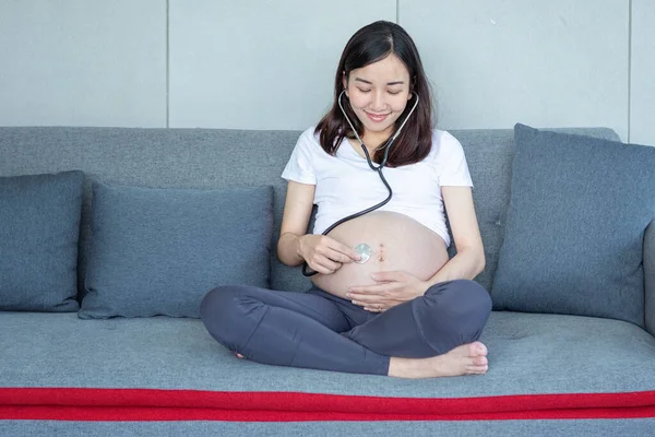 The mother is using stethoscope the doctor listens to the sound of the baby in the stomach. Asian pregnant woman using stethoscope listening her baby on sofa.