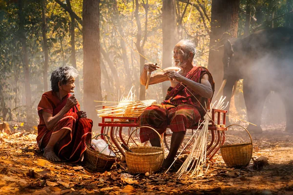 Senior Men and women are weaves basket with a large elephant in the background. Old man and woman weaves bamboo basket or fish trap with elephant in forest. Surin, Thailand