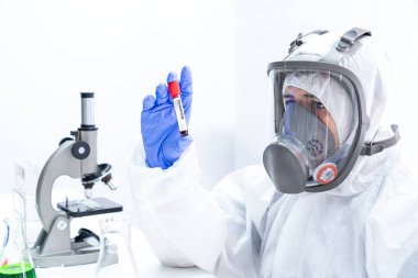 Tehlikeli madde giysisi ve maskeli Asyalı epidemiyoloji uzmanı elinde tüp kanı tutuyor. Coronavirus laboratuvar testi. Doktor ya da bilim adamı kandaki 19 virüsü ya da bakteriyi mikroskopla inceliyor..