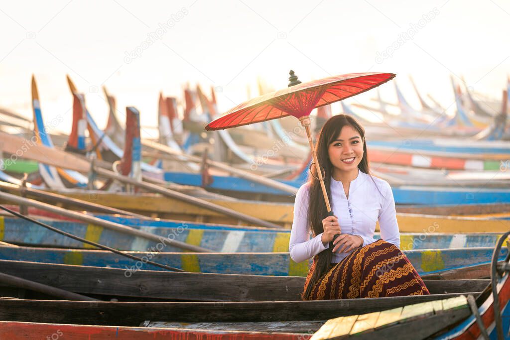 An Asian woman holding a red umbrella sitting on a gondola. A young Burmese woman with a red umbrella on wooden boat at bridge in Asia.U-bein bridge, Mandalay, Myanmar.