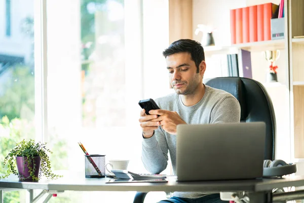 若いビジネスマンが自宅で座って働いています携帯電話やノートパソコンを持っている机の上に — ストック写真