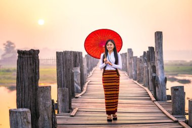 Genç bir Burmalı kadın Asya 'nın en uzun meşe köprüsünde kırmızı şemsiyeyle yürüyor. U-bein köprüsü, Mandalay, Myanmar.