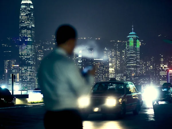 A businessman, at night in Hong Kong, smokes a cigarette, consults his smartphone in front of Hong Kong buildings