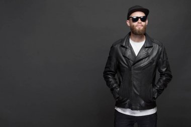 Siyah şapkalı, kot pantolonlu ve güneş gözlüklü deri bisikletçi ceketi veya asimetrik zip ceket giyen adam. Gri arka plan üzerinde Yakışıklı hipster adam