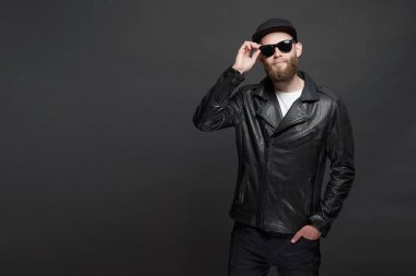 Siyah şapkalı, kot pantolonlu ve güneş gözlüklü deri bisikletçi ceketi veya asimetrik zip ceket giyen adam. Gri arka plan üzerinde Yakışıklı hipster adam