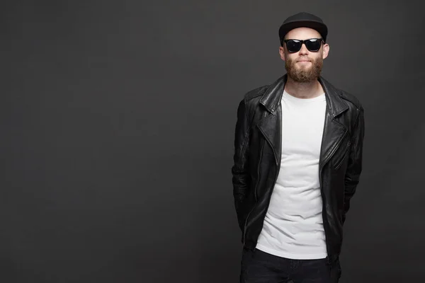 Man wearing leather biker jacket or asymmetric zip jacket with black cap, jeans and sunglasses. Handsome hipster man over gray background — Stock Photo, Image