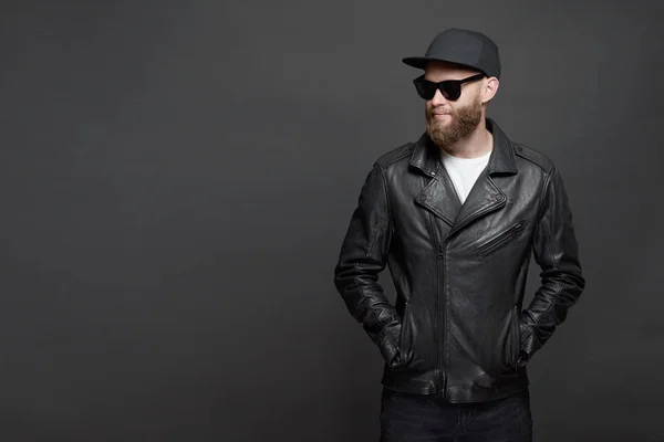 Hombre con chaqueta motorista de cuero o chaqueta con cremallera asimétrica con gorra negra, jeans y gafas de sol. Hombre hipster guapo sobre fondo gris —  Fotos de Stock