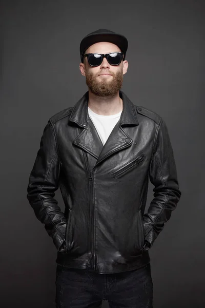Hombre con chaqueta motorista de cuero o chaqueta con cremallera asimétrica con gorra negra, jeans y gafas de sol. Hombre hipster guapo sobre fondo gris —  Fotos de Stock