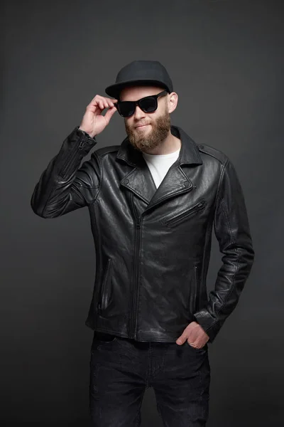 Homem vestindo jaqueta de couro motociclista ou casaco com zíper assimétrico com boné preto, jeans e óculos de sol. Bonito hipster homem sobre fundo cinza — Fotografia de Stock