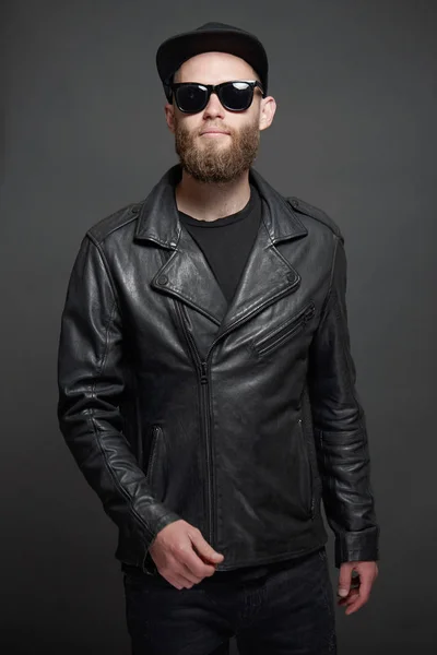 Mann trägt Biker-Lederjacke oder asymmetrische Reißverschlussjacke mit schwarzer Mütze, Jeans und Sonnenbrille. schöner Hipster-Mann mit grauem Hintergrund — Stockfoto